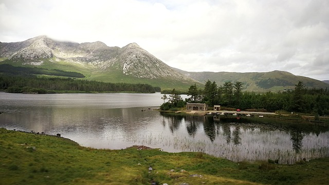 A serene lakeside scene with a small building, surrounded by lush greenery and mountains in the background.