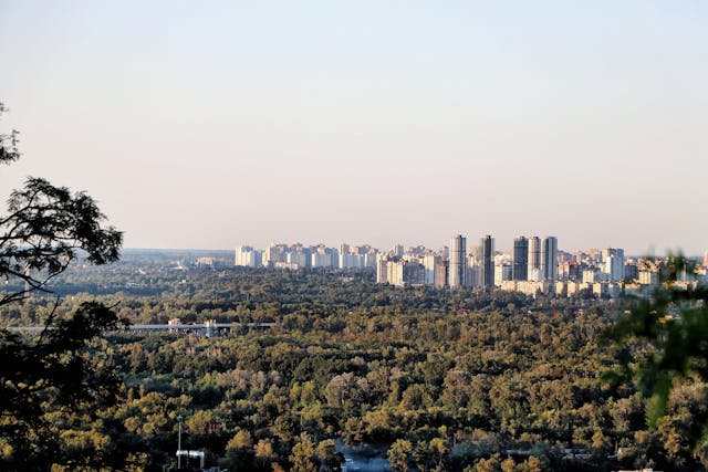 A city skyline with tall buildings rising above a dense forested area under a clear sky.