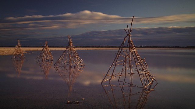 A serene waterscape featuring four triangular wooden structures made of intertwined branches, partially submerged in calm, reflective water.